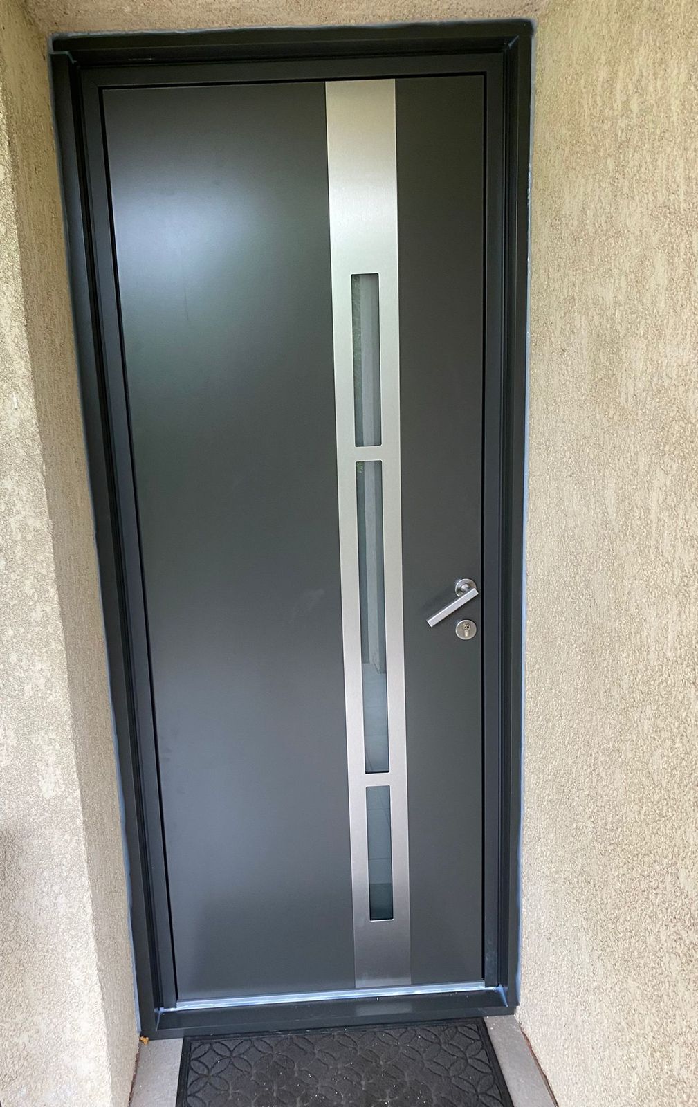 Porte d'entrée gris anthracite avec bande verticale en inox et petits inserts vitrés, sous un porche crépi
