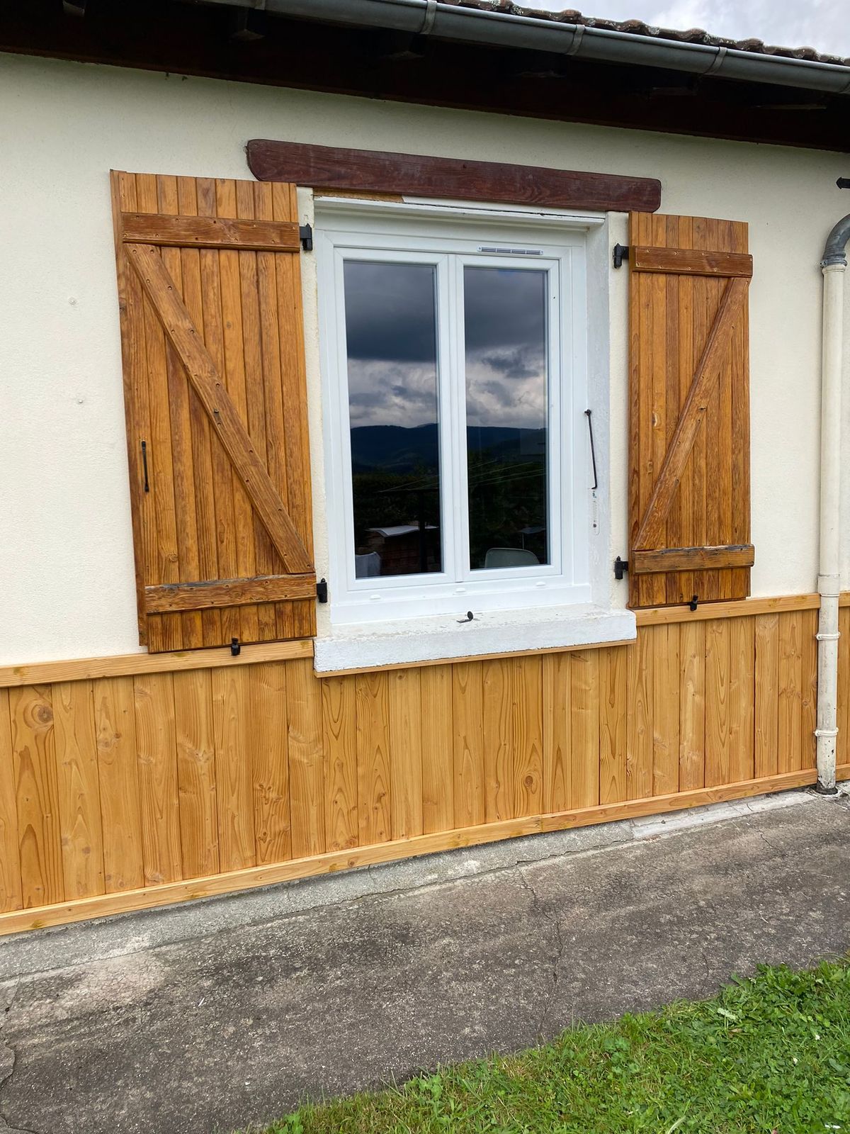 Fenêtre blanche encadrée de volets battants en bois naturel, sur une façade avec soubassement en bardage bois